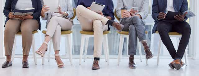 A row of people seated in chairs, some holding books or phones, as if in a waiting room