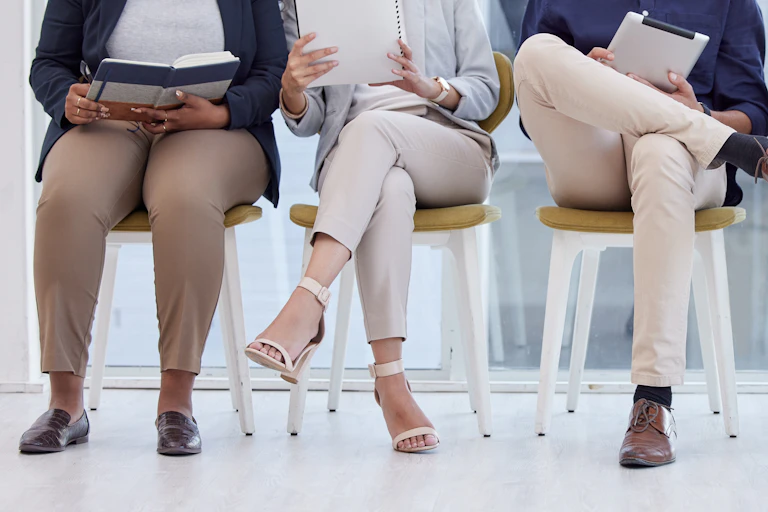 A row of people seated in chairs, some holding books or phones, as if in a waiting room