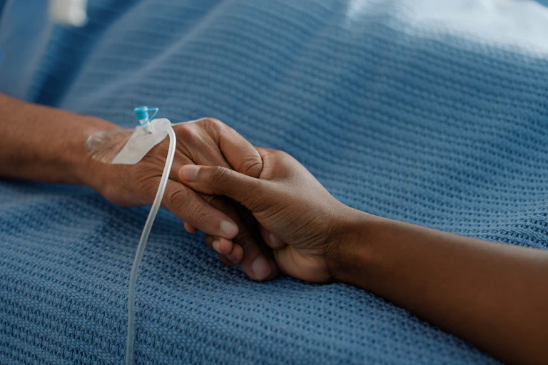 One person's hand holding another person's hand which has a medical drip inserted