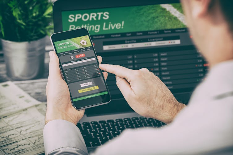 A man holds a phone featuring an online gambling app. There's a laptop in the background also featuring online gambling.