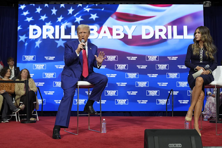 A man with greyish blond hair speaks into a microphone in front of a large screen that reads Drill Baby Drill.
