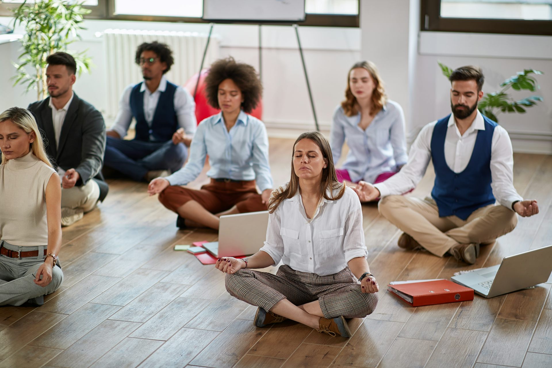 Seven adults in contemporary office-work clothing are sitting cross-legged on a wooden floor in meditative poses.