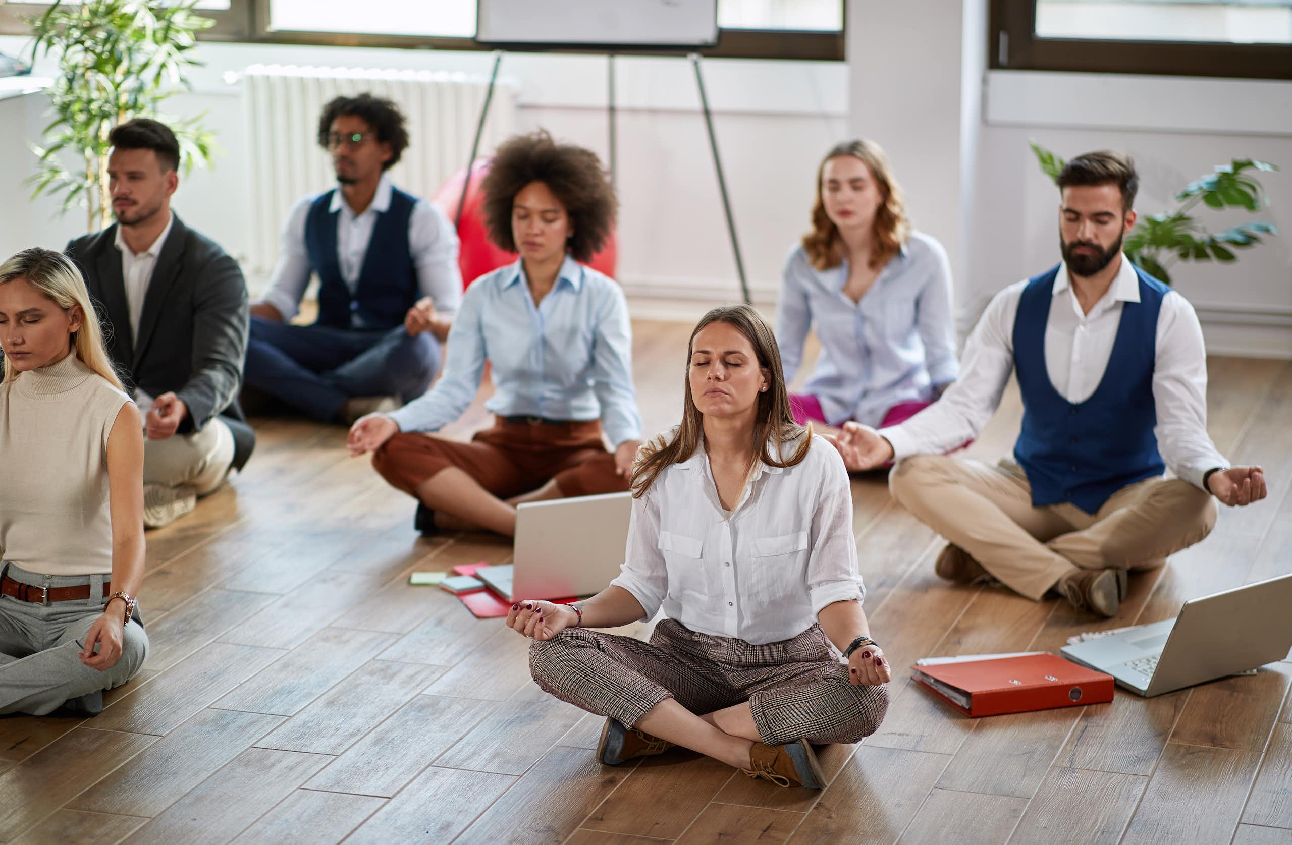 Seven adults in contemporary office-work clothing are sitting cross-legged on a wooden floor in meditative poses.