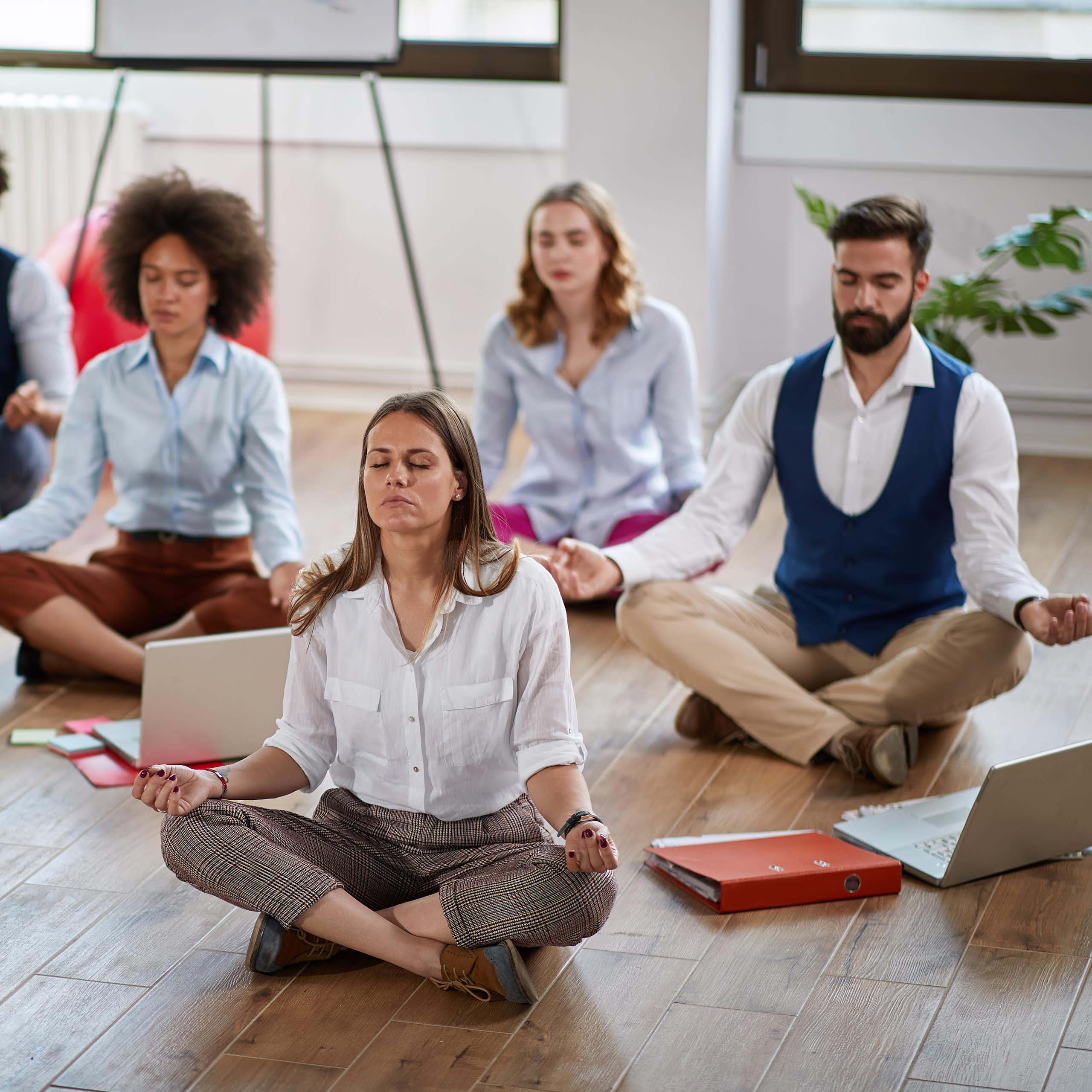 Seven adults in contemporary office-work clothing are sitting cross-legged on a wooden floor in meditative poses.