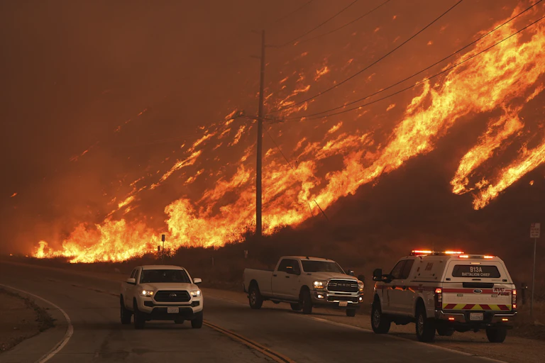 Fire sweeps up a hillside in patches. Vehicles are stopped on the road below.