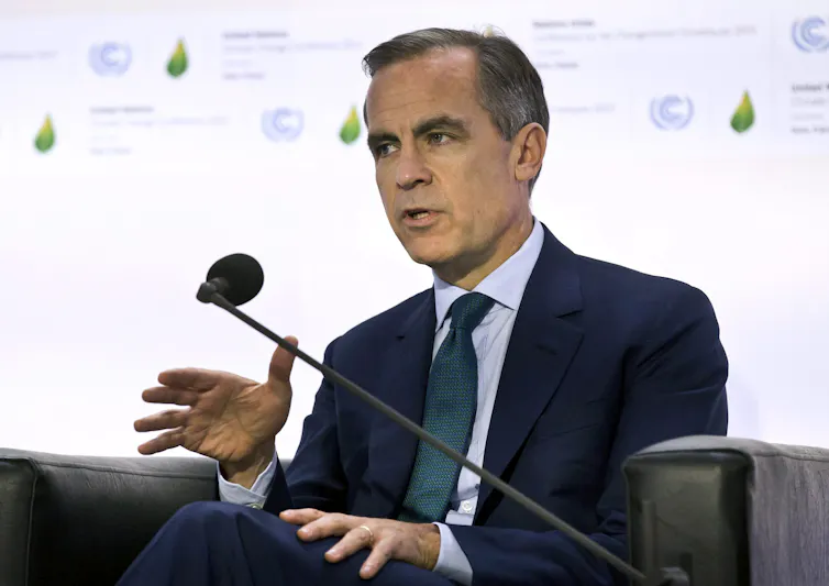 A man in a dark suit gestures as he speaks with a microphone in front of him.