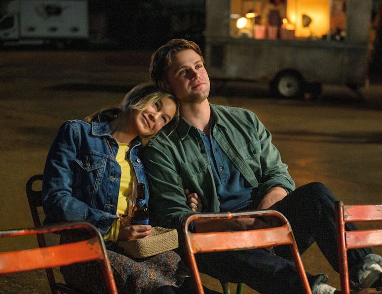 A couple sat on chairs, the older woman has her head on the young man's shoulder.