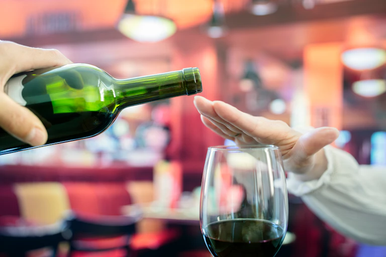 A wine bottle posed over a glass, while a hand covers the glass