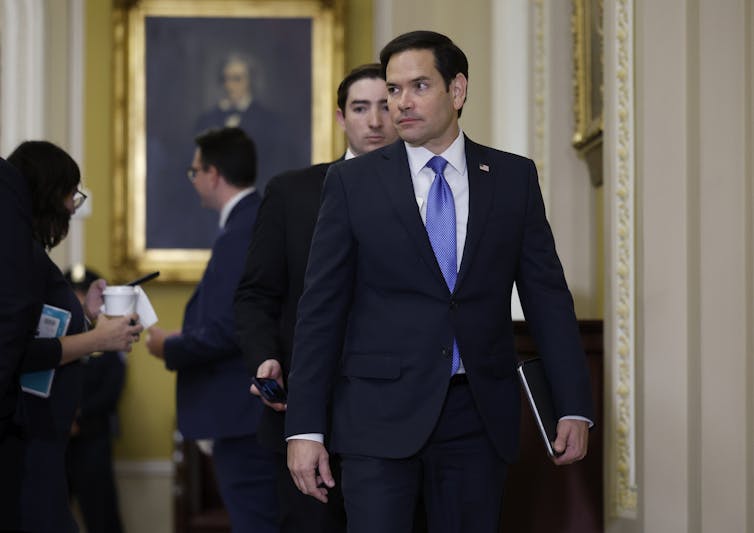 Marco Rubio, standing in a hallway, holding something in his hand.