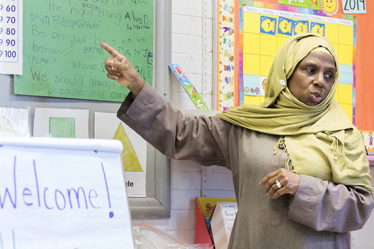 A woman wearing a headscarf stands in front of a teaching chart and points to it