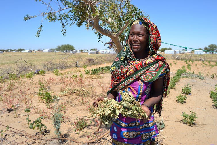 A woman holds two vegetables in her hands while standing in a dry, sparsely populated field with small houses in the distance.