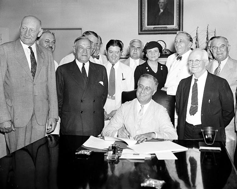 A smiling man in a light-colored suit signs papers at a table, surrounded by about a dozen people.