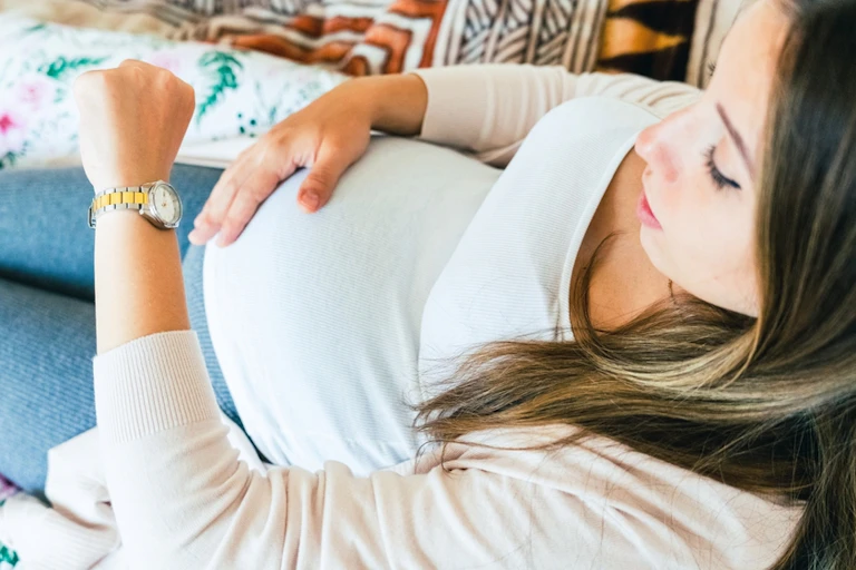 Pregnant woman lying on sofa, looking at watch, one hand on belly