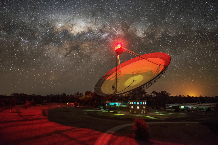 Large, dish-shaped telescope glowing red under a star-filled night sky.