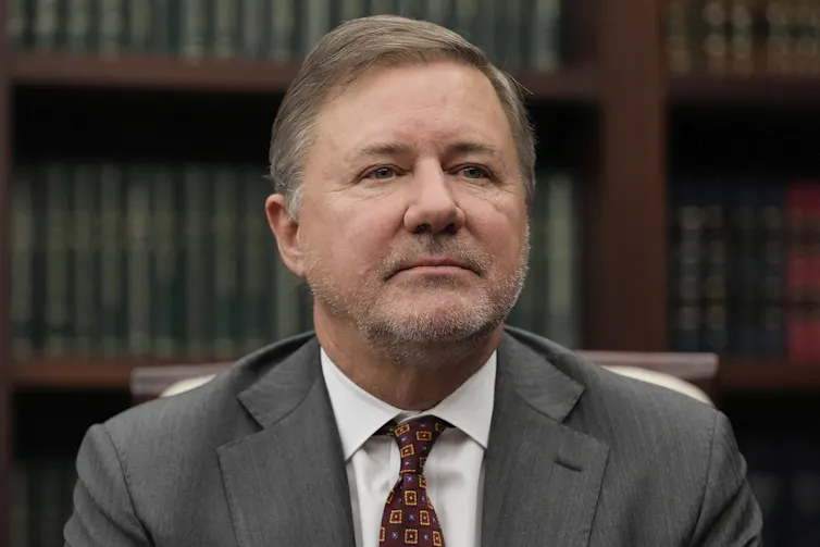 A man in a gray suit and red tie sits with a full bookcase behind him.