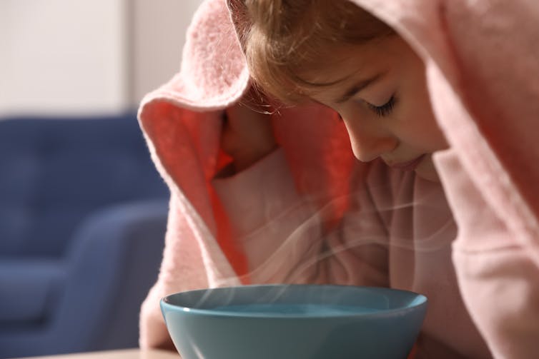 A little girl with a towel over her head inhales steam from a bowl.