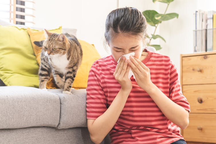 A woman sitting on the floor blows her nose next to a cat.