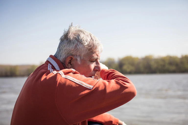 An older man in a red jacket coughs into his elbow.