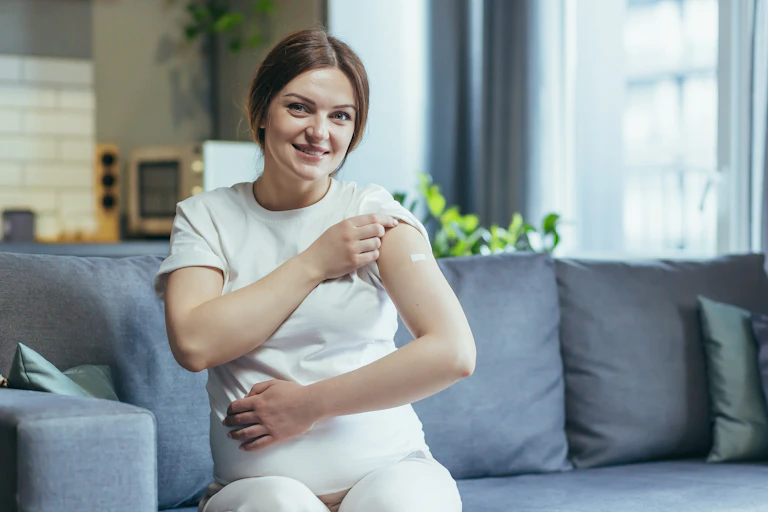 A pregnant woman sitting on a couch revealing a bandaid on her upper arm.