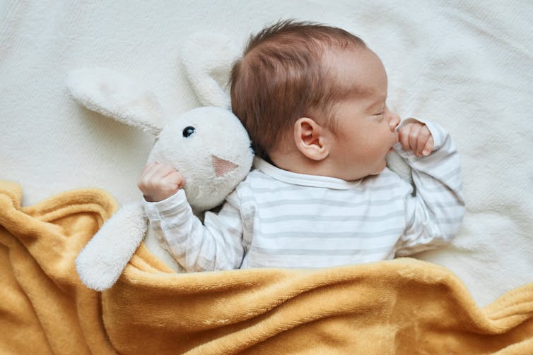 A young baby sleeping under a yellow blanket with a toy bunny.