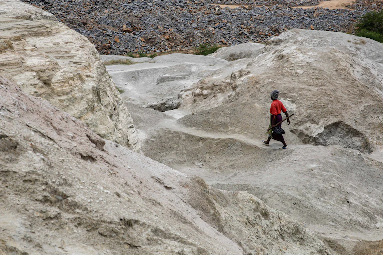 A woman in a red shirt holding a pick and a bag strides across a rocky outcrop.