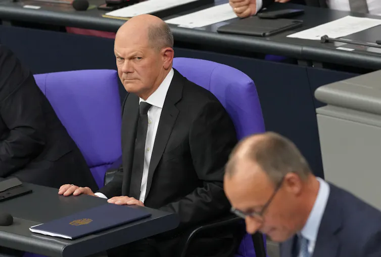 Olaf Scholz scowling in parliament while Friedrich Merz speaks.