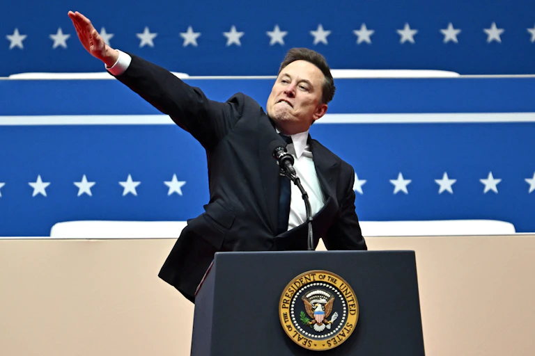 A man stands at a lectern with the seal of the president of the United States and holds his right arm straight out and upward at an angle.