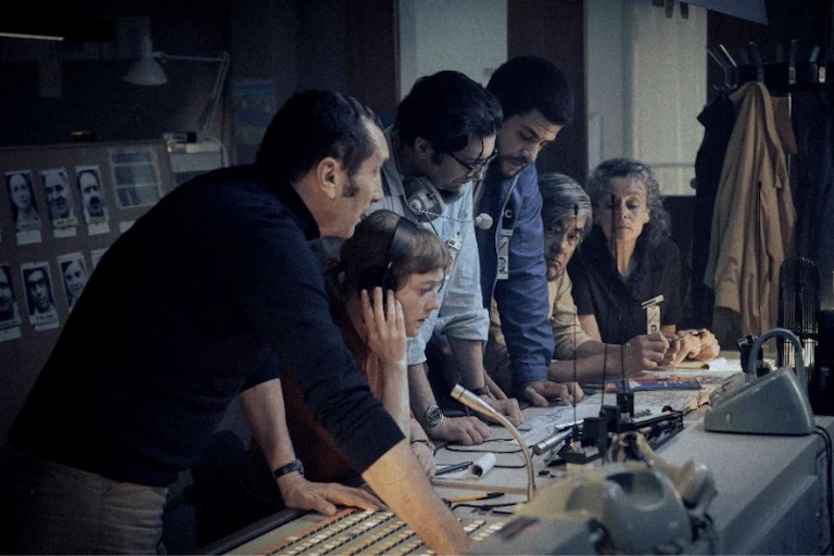 A group of journalists gathered around a broadcast desk.