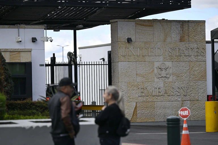 A man and woman stand next to a brick wall with 'United States Embassy' written on it.