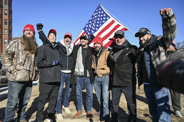 People wearing 'Make America Great Again' hats stand smiling together underneath a U.S. flag.