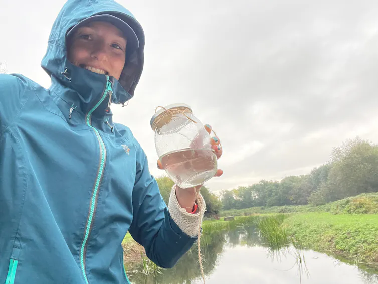 woman in blue raincoat hold jar of water by river
