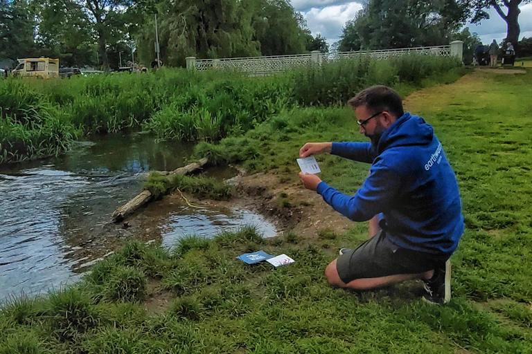 man kneels on ground by river, holding testing equipment