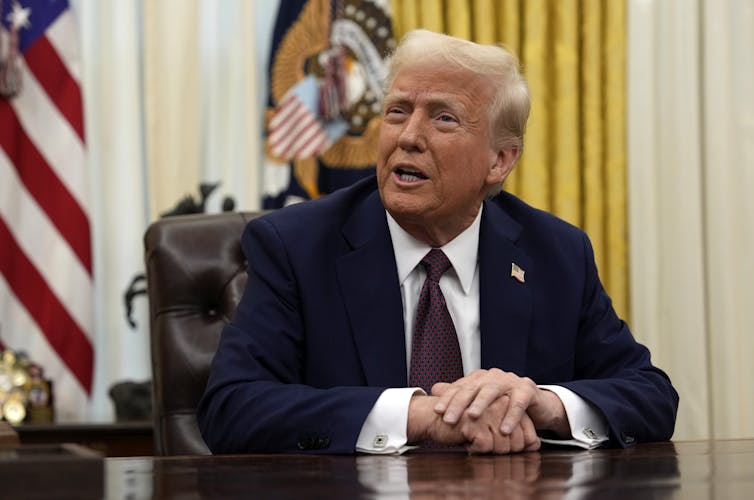 A man with orange-tinged skin and white hair sits behind a desk