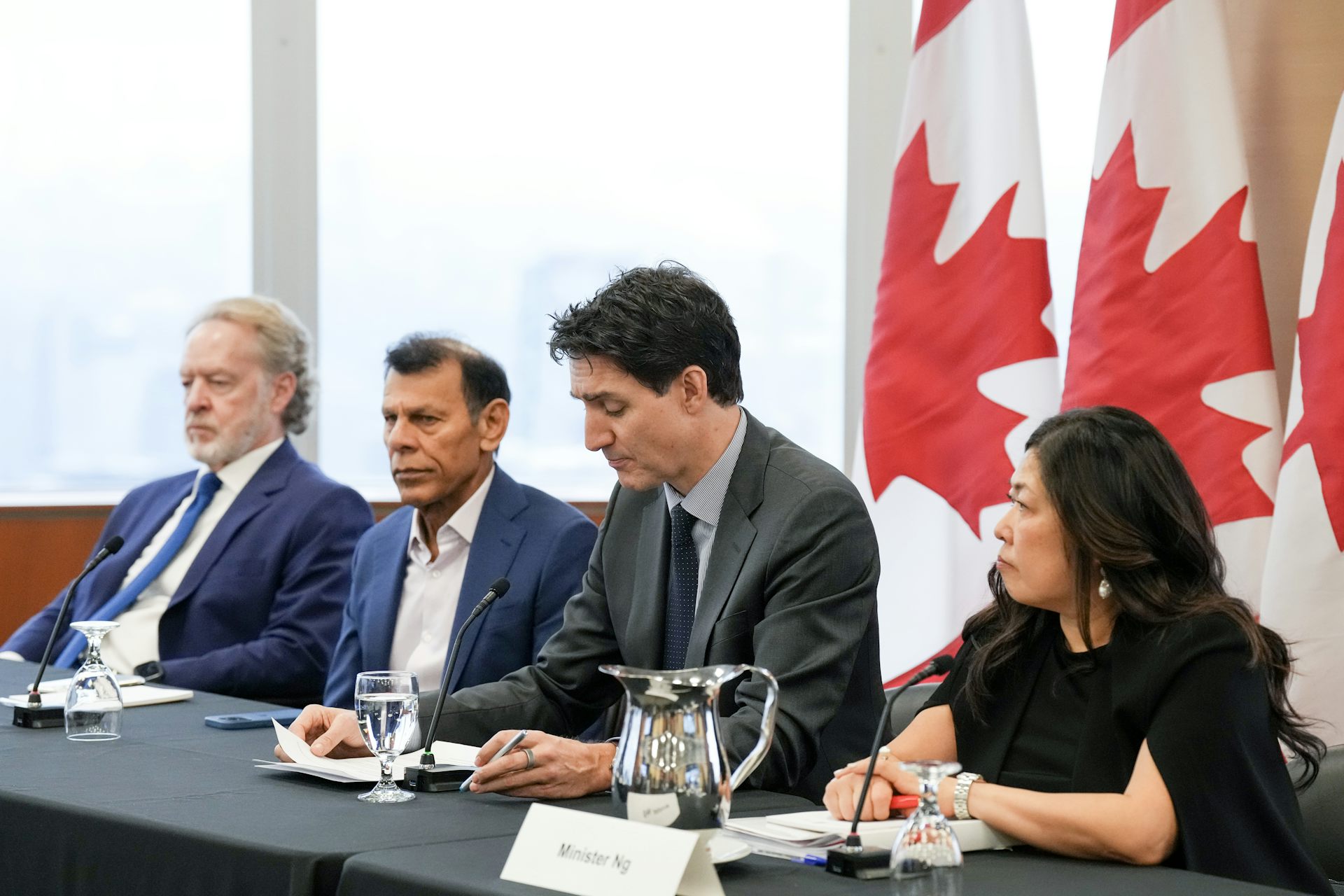 Two white men, a South Asian man, and an East Asian woman sit behind a long table with Canadian flags in the background