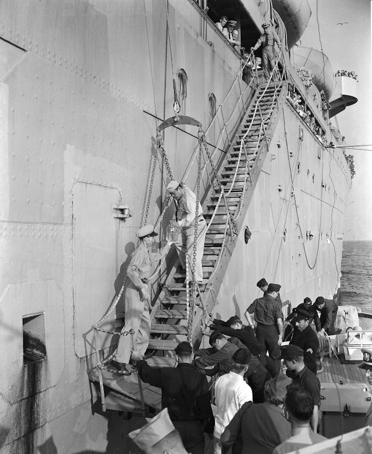 A sailor walking down staircase on side of ship to hand a jar of fluid to a sailor at the bottom, surrounded by other sailors