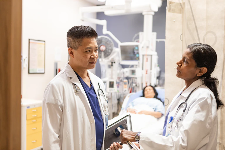 Two physicians look at each other intently with a patient, hooked up to monitors, lying on a bed in the background.