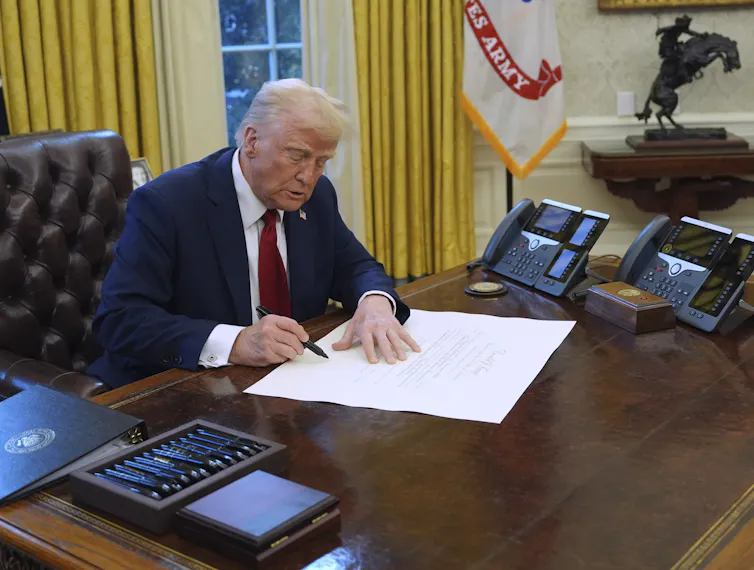 A man with fluffy greyish-blond signs a document at a desk in an ornate office.