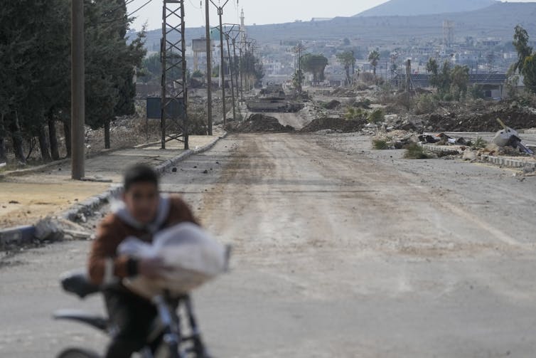 A boy rides a bike on a dusty road with armoured vehicles in the background.