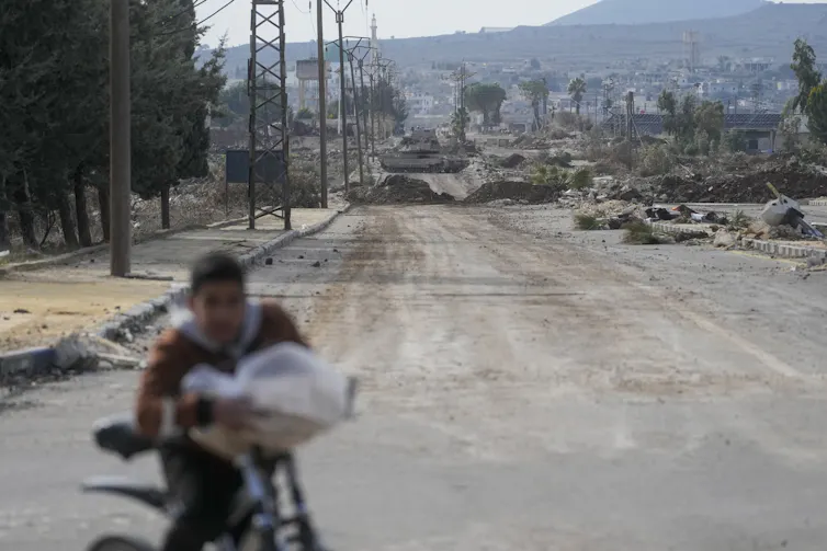 A boy rides a bike on a dusty road with armoured vehicles in the background.