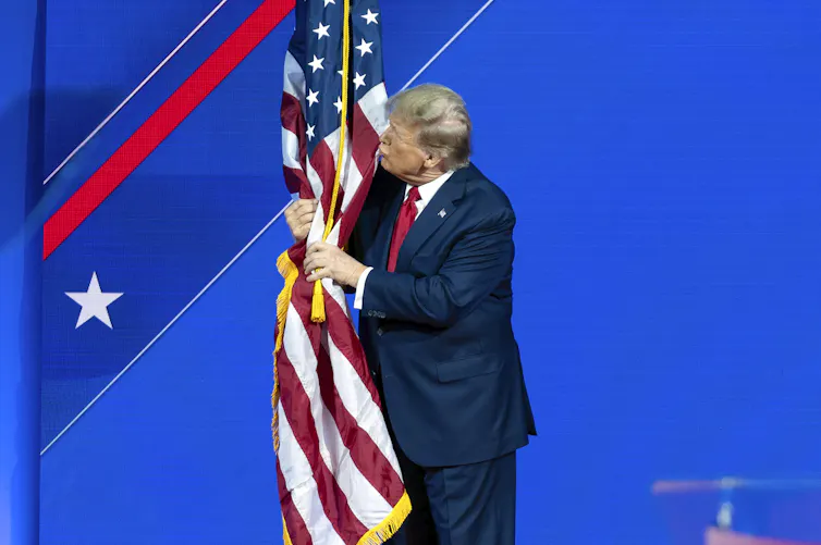 An elderly man with fluffy grey-blond hair kisses an American flag on a stage.