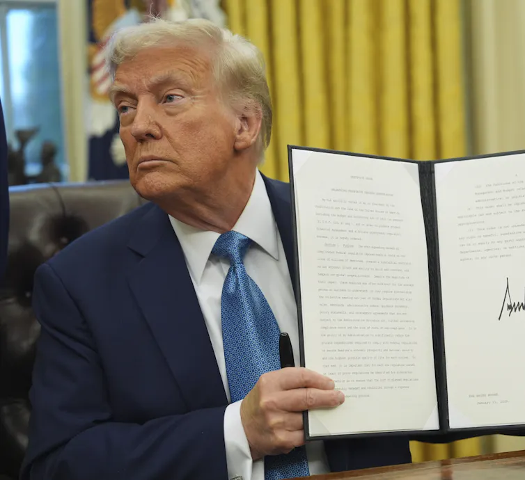 An elderly man with greyish-blond hair holds up a signed document.