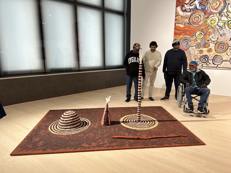 Four men behind artwork in white paint on a red ground.