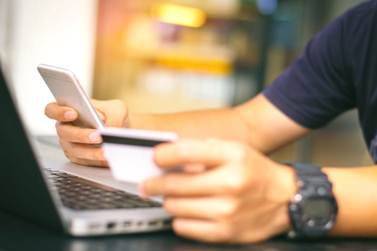 A man sitting at a laptop hold a phone in one hand and a credit card in the other