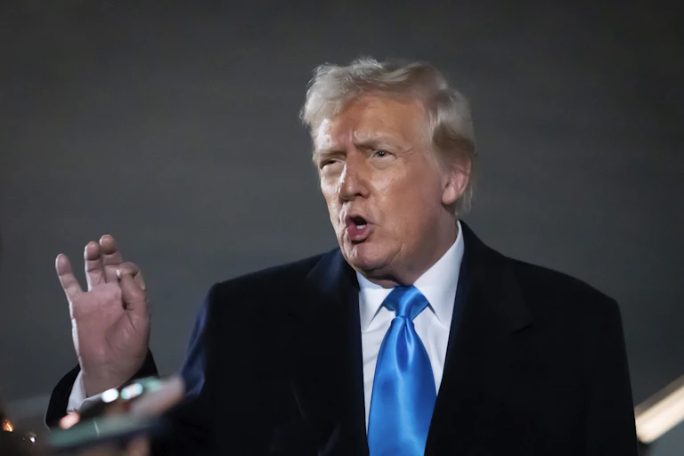 An elderly man with fluffy greyish blond hair holds up his hand as he speaks.