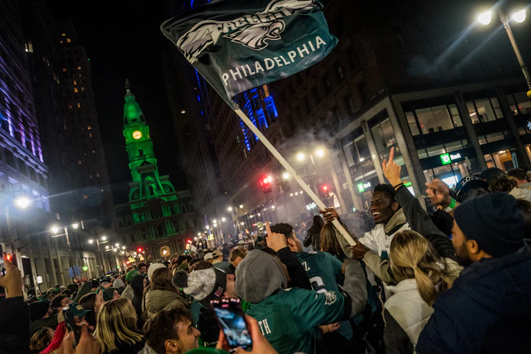 A crowd of people -- many wearing Eagles garb and one waving an Eagles flag -- gather on Broad Street in Philadelphia. Behind them, City Hall is visible, lit in Eagles green.