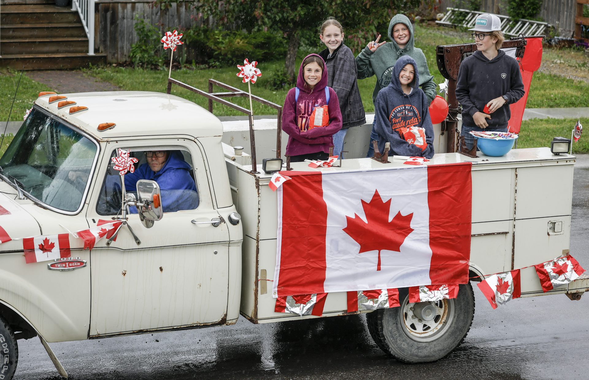 A group of young people smile as they ride in the back of a cream-coloured vintage pickup truck festooned with canadian flags.