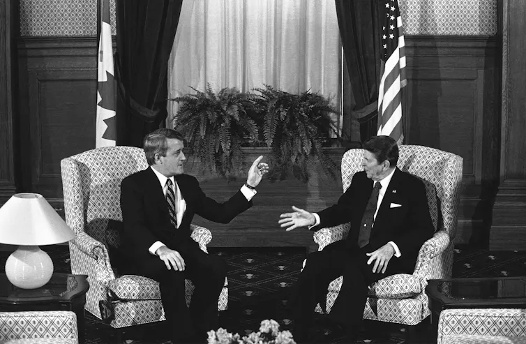 Black and white photo of two white men sitting in armchairs and having a conversation. A Canadian flag stands behind one and and American flag stands behind the other