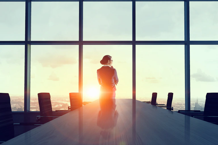 Woman ceo stands at the windows of a boardroom with sun going down