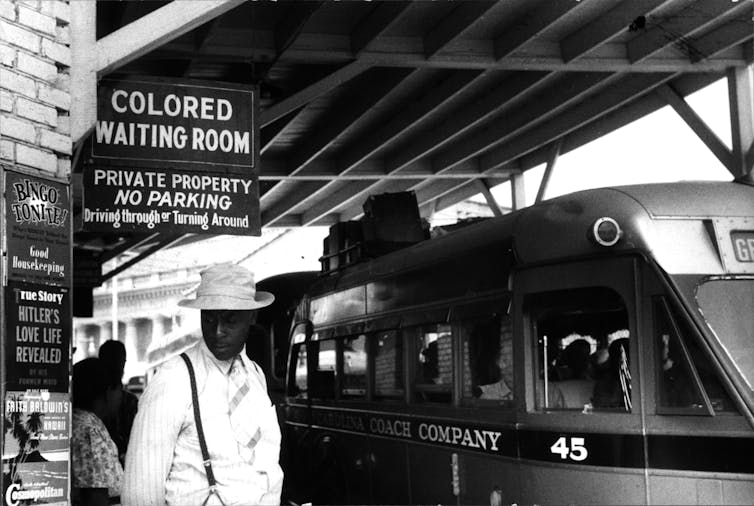 A Black man stands in front of a sign saying Colored Waiting Room at a railway station.
