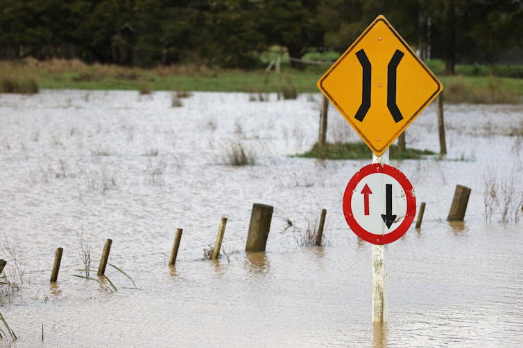 A flooded paddock with a one-way bridge road sign sticking out above the water.